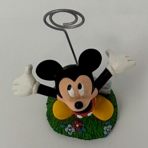 May include: A Mickey Mouse figurine with a silver wire spiral for holding notes or photos. Mickey is in a grassy area with flowers, wearing yellow shoes, a red shirt, and white gloves. The figurine is a decorative item.