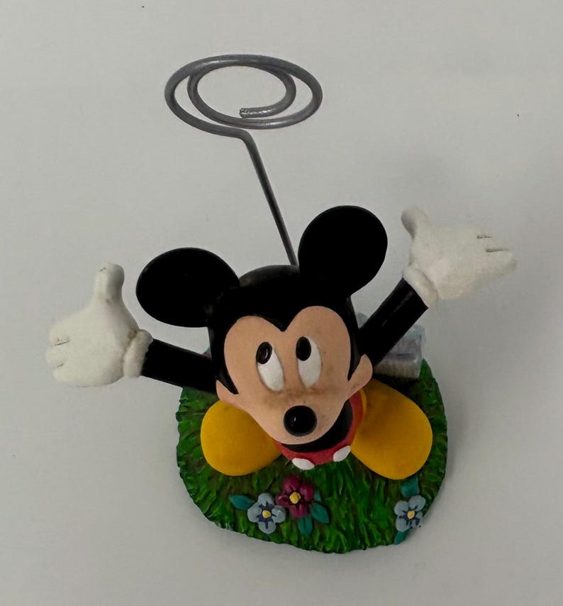 May include: A Mickey Mouse figurine with a silver wire spiral for holding notes or photos. Mickey is in a grassy area with flowers, wearing yellow shoes, a red shirt, and white gloves. The figurine is a decorative item.