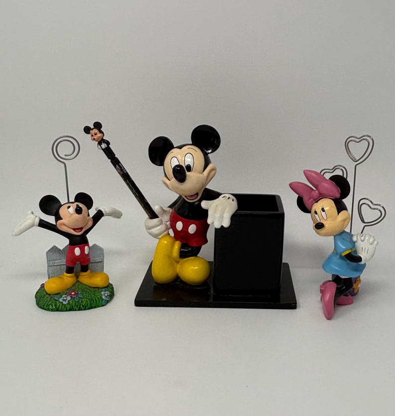May include: Three Disney-themed figurines: Mickey Mouse, Minnie Mouse, and a Mickey Mouse pen holder. Mickey is in a classic pose, Minnie wears a blue dress, and the pen holder is black. The figurines are on a white surface.