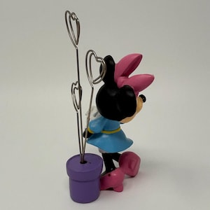 May include: A Minnie Mouse figurine with a light blue dress, pink bow, and heart-shaped wire holders. The figurine is standing next to a purple pot. The base of the figurine is pink.