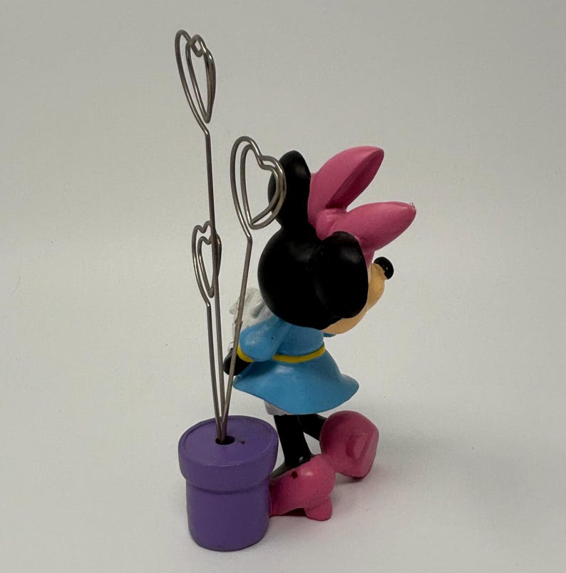 May include: A Minnie Mouse figurine with a light blue dress, pink bow, and heart-shaped wire holders. The figurine is standing next to a purple pot. The base of the figurine is pink.