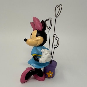 May include: A Minnie Mouse figurine in a blue dress, pink shoes, and a pink bow. The figurine holds a purple pot with heart-shaped wire holders. A yellow flower decorates the pot. The figurine is a decorative item.