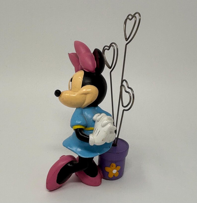 May include: A Minnie Mouse figurine in a blue dress, pink shoes, and a pink bow. The figurine holds a purple pot with heart-shaped wire holders. A yellow flower decorates the pot. The figurine is a decorative item.
