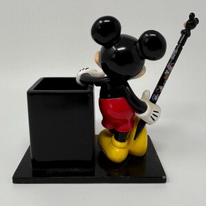 May include: A black Mickey Mouse desk organizer with a pen. The pen holder is a black square, and Mickey is in his classic outfit: red shorts, yellow shoes, and white gloves. The pen is black with colorful Disney character designs.