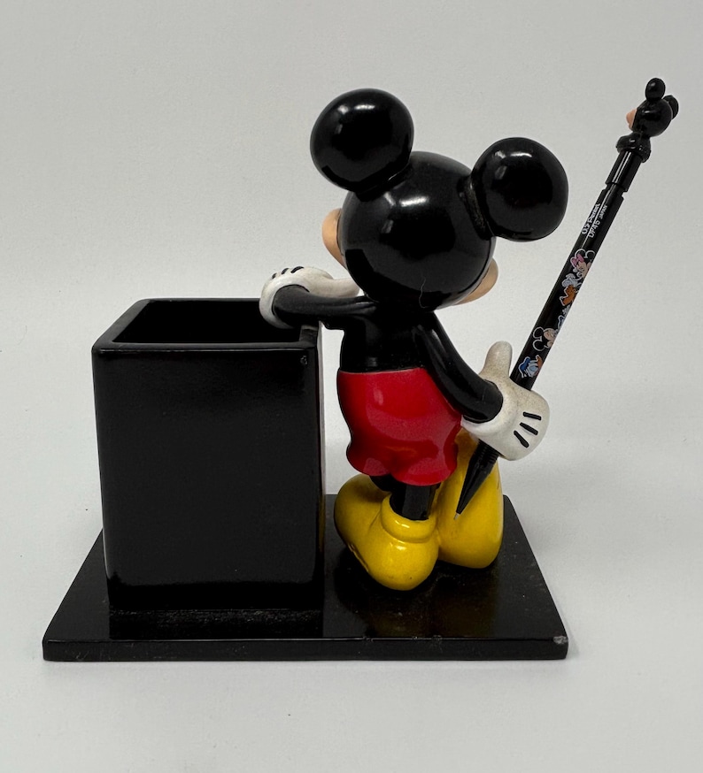 May include: A black Mickey Mouse desk organizer with a pen. The pen holder is a black square, and Mickey is in his classic outfit: red shorts, yellow shoes, and white gloves. The pen is black with colorful Disney character designs.