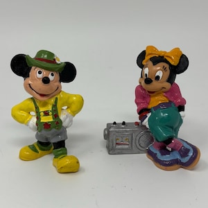 May include: Two painted figurines of cartoon mice. One mouse wears a green hat, yellow shirt, and gray shorts with green suspenders. The other mouse wears a yellow bow, pink top, and teal pants, leaning against a small gray rectangular radio.