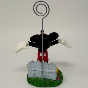 May include: A Mickey Mouse figurine with outstretched arms, wearing red shorts, white gloves, and black ears. The figurine is holding a silver wire with a spiral top, and is standing in front of a light blue picket fence on a green base.