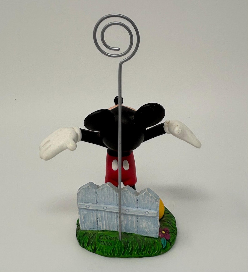 May include: A Mickey Mouse figurine with outstretched arms, wearing red shorts, white gloves, and black ears. The figurine is holding a silver wire with a spiral top, and is standing in front of a light blue picket fence on a green base.