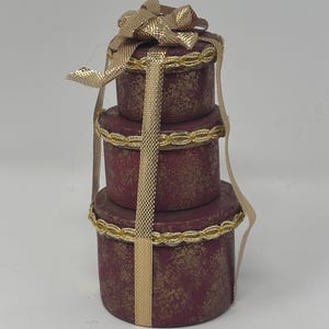 May include: A stack of three round gift boxes in burgundy with gold accents. The boxes are tied with a gold ribbon and bow. The boxes have a gold trim around the lids and bases.