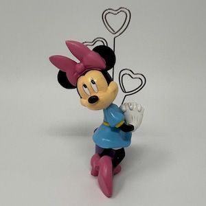 May include: A Minnie Mouse figurine in a blue dress, pink bow, and shoes, holding a white glove. The figurine is paired with a silver heart-shaped memo holder.
