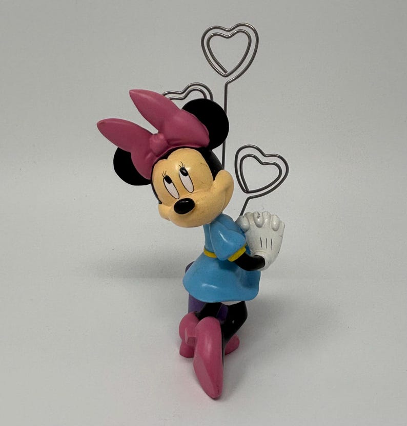 May include: A Minnie Mouse figurine in a blue dress, pink bow, and shoes, holding a white glove. The figurine is paired with a silver heart-shaped memo holder.