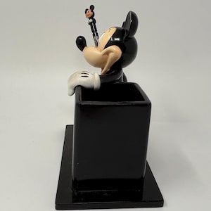 May include: A black Mickey Mouse pen holder with a pen featuring Mickey Mouse on top. The pen holder is rectangular and has a small base. The Mickey Mouse figure is black, white, and tan.