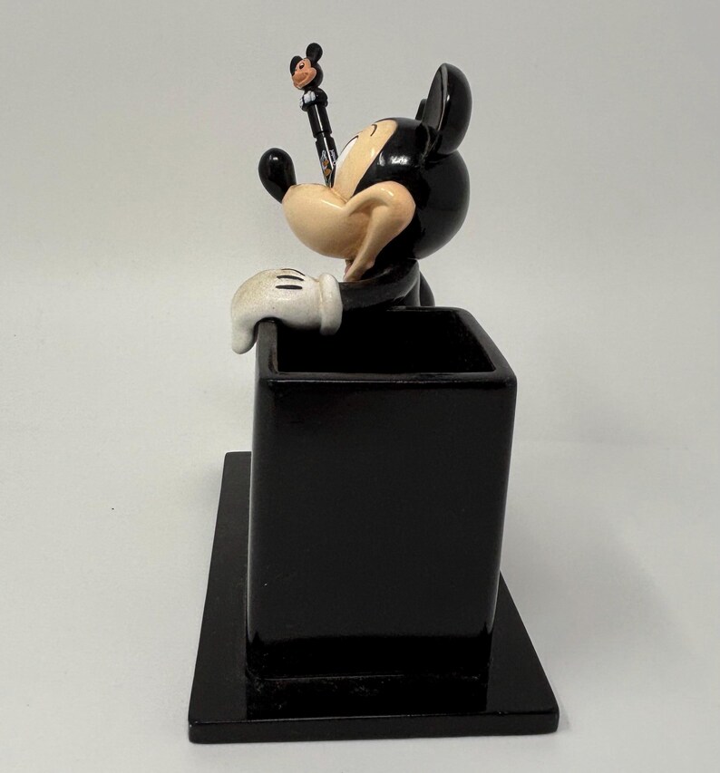 May include: A black Mickey Mouse pen holder with a pen featuring Mickey Mouse on top. The pen holder is rectangular and has a small base. The Mickey Mouse figure is black, white, and tan.