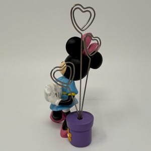 May include: A Minnie Mouse figurine holding a white bag, with three heart-shaped wire holders emerging from a purple flower pot. The figurine wears a blue dress, pink bow, and shoes. The flower pot has a yellow flower.