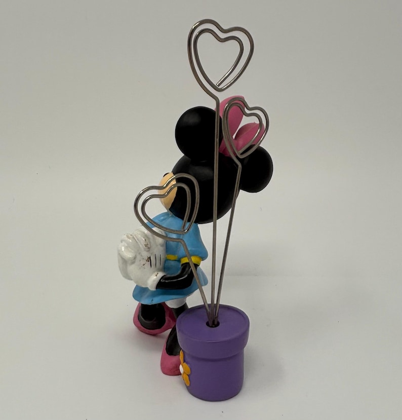 May include: A Minnie Mouse figurine holding a white bag, with three heart-shaped wire holders emerging from a purple flower pot. The figurine wears a blue dress, pink bow, and shoes. The flower pot has a yellow flower.