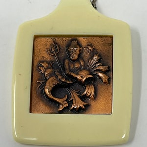 May include: A white plastic pendant with a copper-colored relief of a mythical creature holding a trident. The creature has a beard and is surrounded by fish and seaweed.