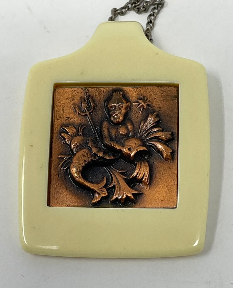 May include: A white plastic pendant with a copper-colored relief of a mythical creature holding a trident. The creature has a beard and is surrounded by fish and seaweed.