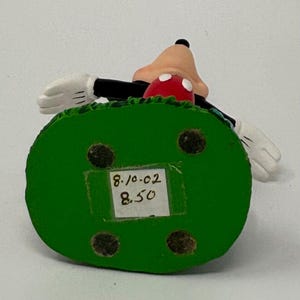 May include: A Mickey Mouse figurine with a green base. The figurine features Mickey's face, red shorts, and white gloves. The base has a sticker with the numbers "8.10.02" and "8.50".