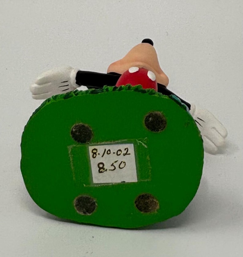 May include: A Mickey Mouse figurine with a green base. The figurine features Mickey's face, red shorts, and white gloves. The base has a sticker with the numbers "8.10.02" and "8.50".