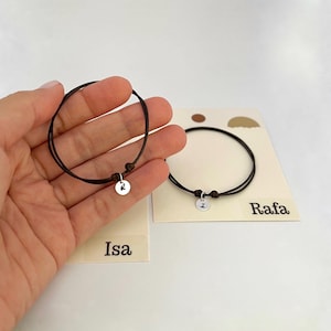 Pode incluir: Duas pulseiras de cordão castanho escuro, cada uma com um pequeno pendente redondo de metal prateado estampado com uma letra preta. Um pendente apresenta a letra R e o outro a letra I. As pulseiras são apresentadas em cartões com as etiquetas Isa e Rafa.