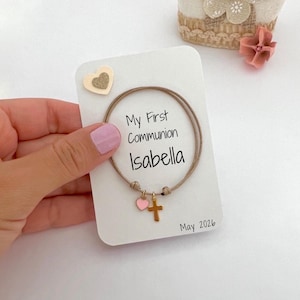 Könnte beinhalten: Ein beiges Kordelarmband mit einem kleinen goldenen Kreuzanhänger und einem rosa Herzanhänger, präsentiert auf einer weißen Karte mit dem Text My First Communion, Isabella und May 2026 sowie einem kleinen dekorativen Herz oben links.