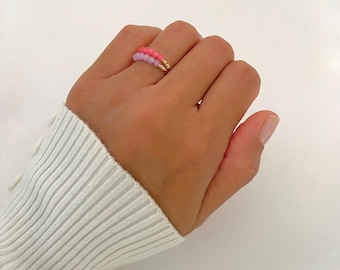 Peut inclure: Une bague à double rangée composée d'une ligne de petites perles rondes rose vif et d'une ligne de petites perles rondes violet clair. Les perles sont enfilées sur un fil métallique doré fin qui entoure le doigt en deux bandes distinctes.