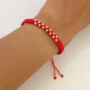 Pode incluir: Uma pulseira de cordão vermelho com uma fila central de pequenas contas esféricas douradas e polidas tecidas no fio. A pulseira tem duas pontas de cordão vermelho finas e pendentes, cada uma terminada com uma pequena conta dourada na ponta.