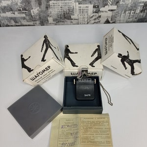 May include: A black pedometer with a silver chain and a black leather case. The case has the word "ЗАРЯ" printed on it. The pedometer is in a box with the word "ШАГОМЕР" printed on it. The box is white with a black silhouette of a person walking on it.