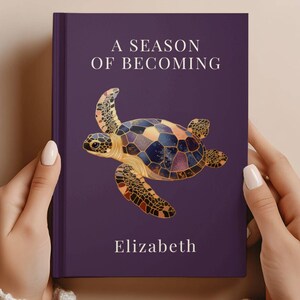 May include: A purple book cover with the title "A Season of Becoming" and the name "Elizabeth" in white text. A colorful turtle illustration is centered on the cover. The book is held by two hands.