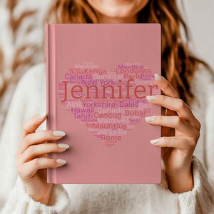 May include: A pink journal with the name "Jennifer" in a heart-shaped design. The heart is filled with names of places like Paris, London, and Hawaii. The journal is held by a person.