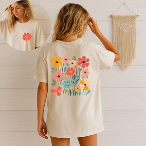 May include: Cream-colored t-shirt featuring a colorful floral design on the back and a smaller flower on the front. The graphic includes various flowers in shades of yellow, pink, red, and blue, creating a cheerful and vibrant look.