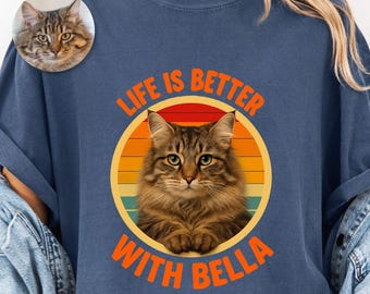 Pode incluir: Uma t-shirt azul-marinho com um gráfico circular com um fundo de pôr do sol em riscas laranja, amarelas e vermelhas. Um gato malhado castanho está centrado no círculo com o texto laranja "LIFE IS BETTER WITH BELLA" arqueado à volta.