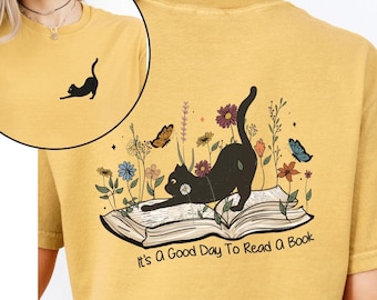 Könnte beinhalten: Ein senfgelbes T-Shirt mit einem schwarzen, sich streckenden Katzenmotiv auf einem offenen Buch, umgeben von bunten Wildblumen und Schmetterlingen. Der Text "It's A Good Day To Read A Book" steht unter dem Bild. Vorne ist eine kleine Katze.