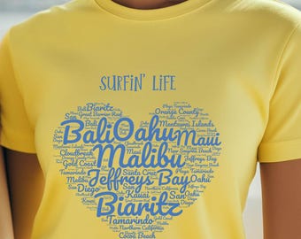 Puede incluir: Una camiseta de manga corta de color amarillo brillante que presenta el texto SURFIN' LIFE sobre un gran gráfico en forma de corazón compuesto por varios nombres azules de lugares de surf famosos como Bali, Oahu, Malibu y Biarritz.