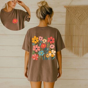 May include: A brown t-shirt featuring a colorful floral design on the back and a single flower on the front. The flowers are in shades of yellow, pink, red, and blue. The shirt has short sleeves and a relaxed fit.