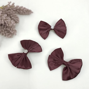 Puede incluir: Tres lazos de pelo de satén burdeos con diferentes adornos. Un lazo tiene una perla, otro tiene un strass y el último tiene un strass transparente.