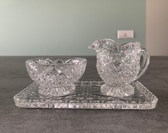 May include: A clear glass sugar and creamer set with a tray. The set has a diamond pattern and is on a gray surface.