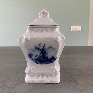 Op de afbeelding: Witte porseleinen pot met een blauw-wit windmolenontwerp. De pot heeft een deksel met een handvat.