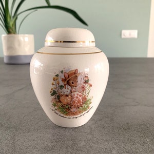 Könnte beinhalten: Eine weiße Keramikdose mit goldenem Deckel und einem Blumendesign mit einem kleinen Tier, das einen Hut trägt. Die Dose hat den Text "A little kindness can bring a lot of happiness" auf dem Boden aufgedruckt.