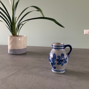 Op de afbeelding: Een kleine blauwe en witte keramische kan met een bloemenmotief. De kan heeft een handvat en een rond lichaam.