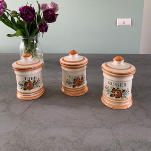 May include: Three ceramic canisters with orange and white floral designs. The canisters are labelled "Thee", "Koffie", and "Suiker".
