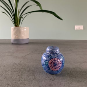 Op de afbeelding: Een kleine blauwe en witte porseleinen pot met een bloemenmotief. De pot heeft een deksel en is versierd met roze en groene bloemen.