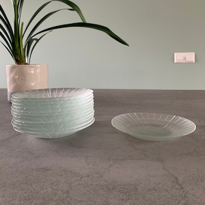 Pode incluir: Uma pilha de dez pratos de vidro transparente com um padrão radial sobre uma bancada cinza. Os pratos estão empilhados em uma pilha organizada, com o prato superior ligeiramente deslocado dos outros. Um único prato é colocado na bancada à direita da pilha.