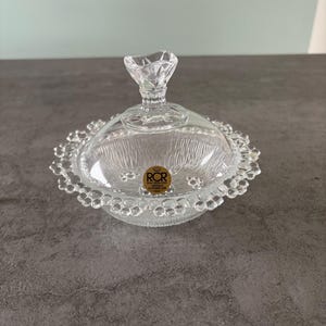 May include: A clear glass candy dish with a lid. The dish has a textured interior and a decorative, scalloped edge. The lid features a small handle. A gold sticker with text is visible on the dish.