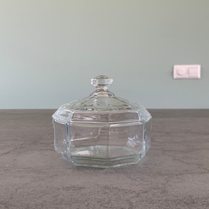 Könnte beinhalten: Ein klares Glasgefäß mit Deckel. Das Gefäß hat eine sechseckige Form und ist aus klarem Glas gefertigt. Der Deckel ist ebenfalls aus klarem Glas und hat einen kleinen Knopf oben.