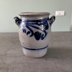 Könnte beinhalten: Eine kleine, graue Steinzeug-Vase mit zwei Henkeln. Die Vase hat ein blaues und weißes Blumenmuster.