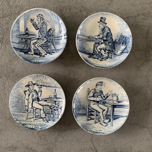 May include: Set of four blue and white ceramic plates with illustrations of men drinking. Each plate features a different drink and the text "Le ..." below the image. The plates are approximately 6 inches in diameter.