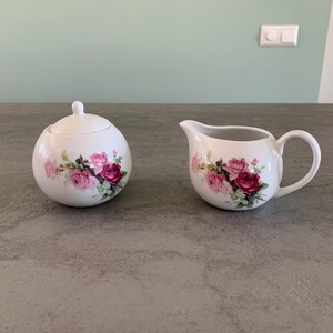 Può includere: Un set di zuccheriera e lattiera bianca con un motivo floreale con rose rosa e foglie verdi. Il set è in porcellana e ha una finitura lucida.