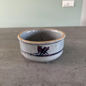 Puede incluir: Un pequeño cuenco de cerámica gris con un borde marrón claro y un diseño morado en el lateral.