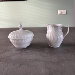 Può includere: Una zuccheriera in porcellana bianca con coperchio e una piccola lattiera. Entrambi i pezzi presentano un design a spirale a coste. La zuccheriera ha una maniglia decorativa sul coperchio. La lattiera ha un manico e un bordo smerlato.